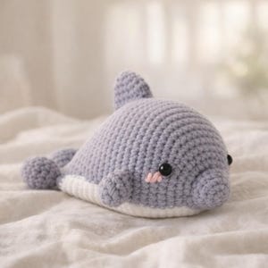May include: A handmade crochet dolphin in shades of gray and white. The plush toy features a rounded body, a dorsal fin, and small flipper-like appendages. It has black safety eyes and a pink blush on its cheeks.