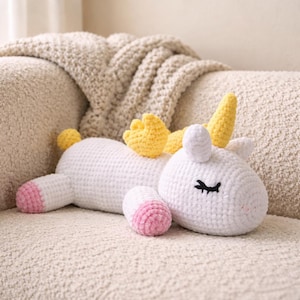 May include: A white crocheted unicorn plush toy with yellow horn and mane details, and pink hooves. The unicorn is lying down on a cream-colored sofa, with a beige knitted throw in the background. The toy is soft and cuddly.