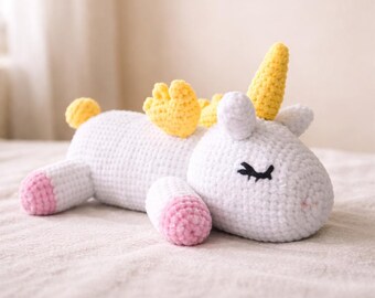 May include: A white crocheted unicorn plush toy with yellow horn, mane, and tail accents. The unicorn has pink hooves and closed black eyes. The toy is lying down on a light-colored surface.