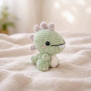 Op de afbeelding: Een klein, gehaakt dinosaurus speelgoed in groen- en wittinten. De dinosaurus heeft een vriendelijke uitdrukking met een zwart oog, een gestikte glimlach en roze wangen. Het heeft grijze stekels op de rug en zit op een zacht, crèmekleurig oppervlak.