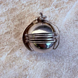 May include: A silver-toned metal locket pendant shaped like a sphere. The locket features a hinged mechanism with multiple horizontal segments that can expand outward, connected by small circular rivets on the sides and a top bail loop.