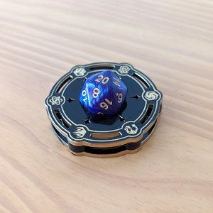 May include: A blue and gold dice tower with a blue 20-sided die resting on top. The die has the numbers 10, 8, 16, 9, 4, and 20 visible.