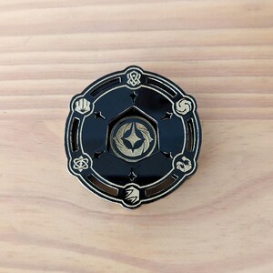 May include: A black and gold circular pin with a starburst design in the center. The pin has a decorative border with eight different symbols around the edge.