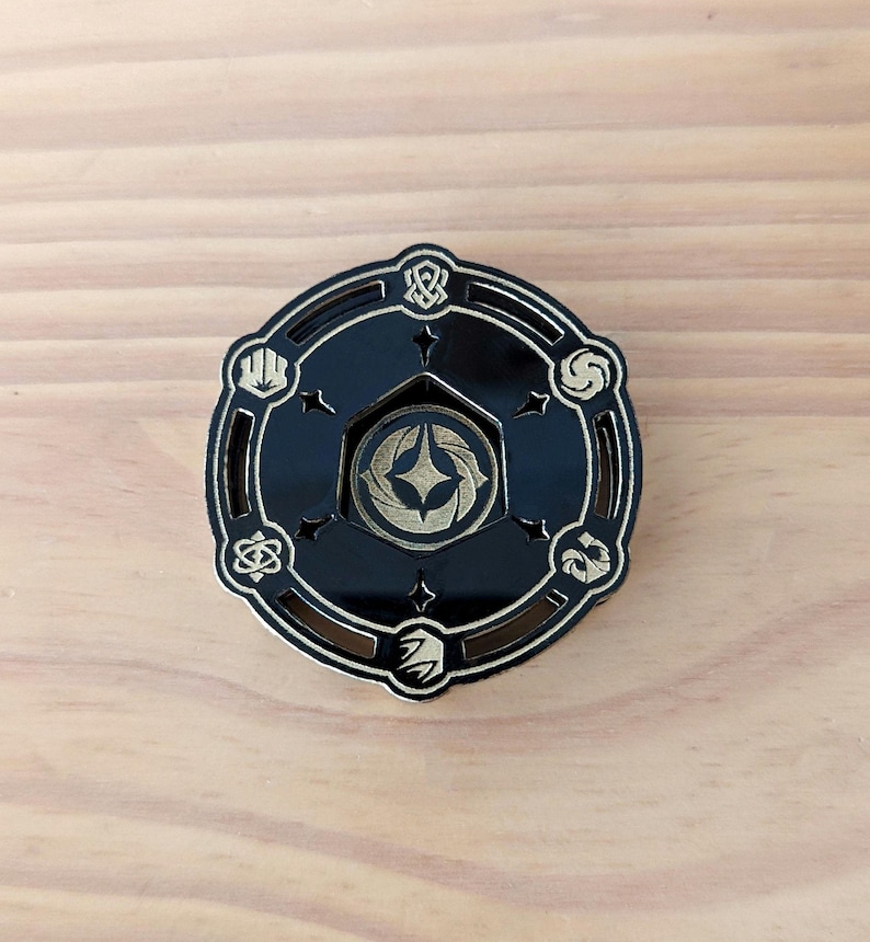 May include: A black and gold circular pin with a starburst design in the center. The pin has a decorative border with eight different symbols around the edge.