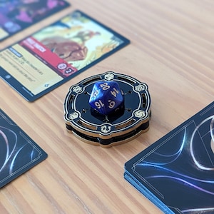 May include: A blue 20-sided die with gold numbers sits on a black and gold wooden dice tower. The tower has a circular base and a raised platform with a design of five symbols.