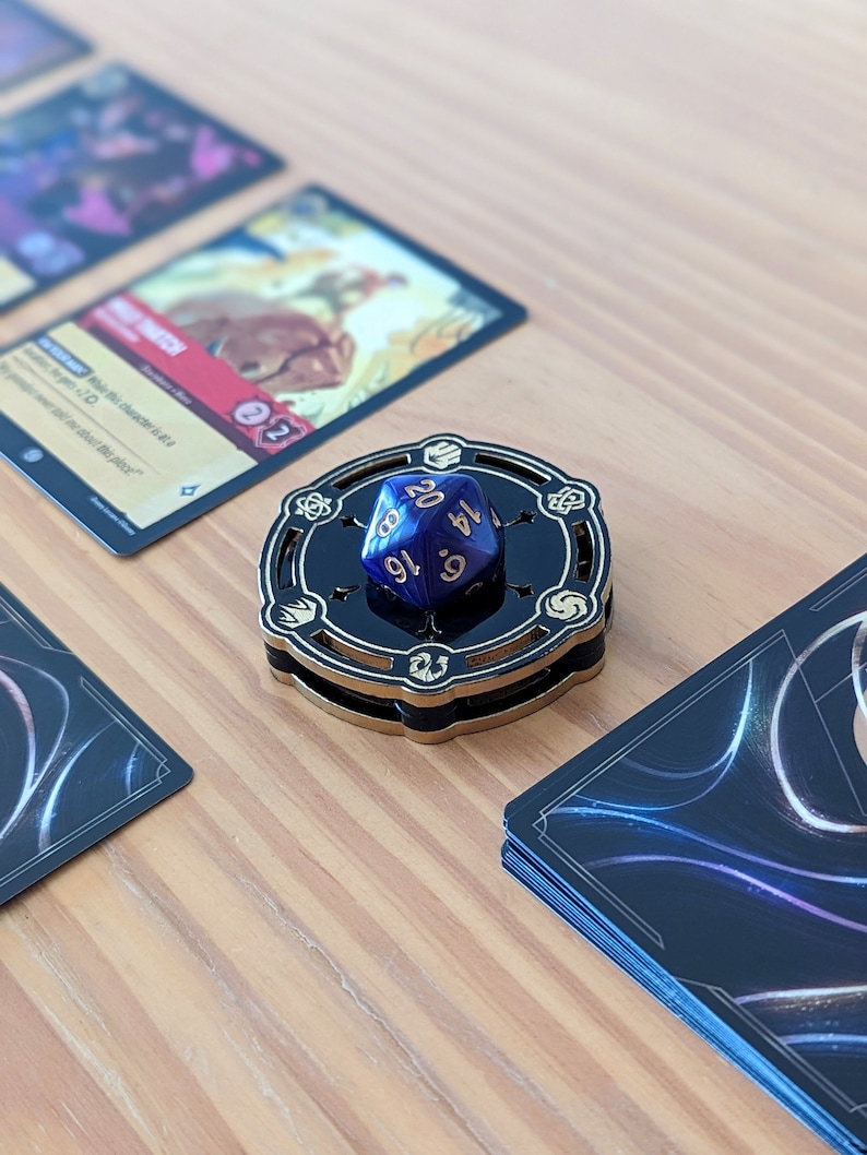 May include: A blue 20-sided die with gold numbers sits on a black and gold wooden dice tower. The tower has a circular base and a raised platform with a design of five symbols.