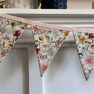 May include: A floral bunting with a pink trim and a white background. The bunting is made of fabric and has a variety of flowers in shades of pink, red, yellow, and green.