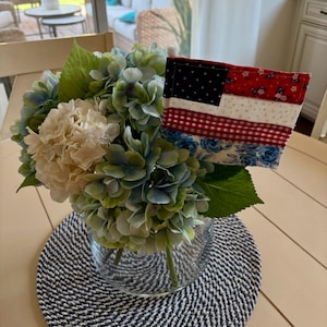 May include: A clear glass vase filled with blue, green, and cream-colored hydrangea flowers and a fabric American flag. The flag features a dark blue canton with white stars, red floral fabric, white star-patterned fabric, and red gingham stripes.
