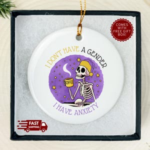 May include: White ceramic ornament with a skeleton illustration holding a coffee mug, wearing a yellow hat, and the text "I don't have a gender, I have anxiety." Comes in a gift box. Gold string for hanging.