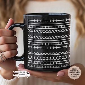 May include: Black ceramic mug with a white geometric pattern. The design features various horizontal lines with shapes like squares, zigzags, and curves. The mug is held in a person's hands.