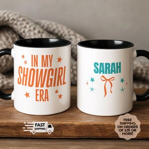 May include: Two white ceramic mugs with black handles and rims. One mug reads "IN MY SHOWGIRL ERA" in orange, with star accents. The other mug says "SARAH" in teal, with a bow and star accents.