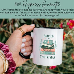 May include: White ceramic mug with a black rim and handle. The mug has a design of a stack of books with the text "Booked for Christmas" and "Spicy Tales" on the books. The mug is being held by a hand.