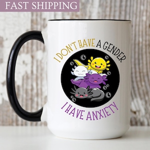 May include: White ceramic mug with a black handle. The mug has a black circle with a white background and a graphic of four axolotls stacked on top of each other. The text "I don't have a gender I have anxiety" is written in a gold font around the circle.