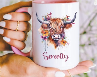 May include: White ceramic mug with a pink rim and handle. The mug features a watercolor illustration of a Highland cow with flowers and butterflies. The text "Serenity" is printed in pink below the image.