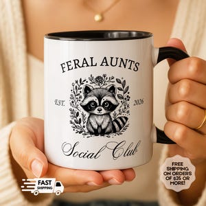 May include: White ceramic mug with a black handle and rim, featuring the text "FERAL AUNTS Social Club" and a raccoon illustration. The mug also includes the text "EST. 2026".
