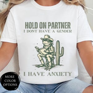 May include: White t-shirt with a green frog wearing a cowboy hat and holding a book. The text on the shirt reads "Hold on partner I don't have a gender I have anxiety".