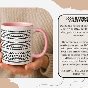 K&ouml;nnte beinhalten: Wei&szlig;e Keramiktasse mit rosa Innenraum und Henkel, verziert mit schwarzen geometrischen Mustern. Die Tasse wird von einer Person in den H&auml;nden gehalten. Die Tasse hat den Text "100% HAPPINESS GUARANTEE".