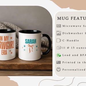 May include: Two white ceramic mugs with black handles and rims. One mug has the text "IN MY SHOWGIRL ERA" in orange, and the other says "SARAH" in teal with a bow graphic. Both mugs are on a wooden surface.