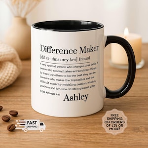 May include: White ceramic mug with a black interior and handle. The mug features the text "Difference Maker" with a definition and the name "Ashley". The mug is on a wooden surface with coffee beans and a lit candle.