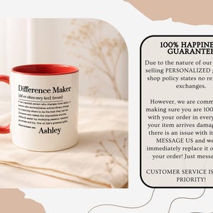 May include: A white ceramic mug with a red interior and handle. The mug features the text "Difference Maker" with a definition and the name "Ashley". The mug is on a white cloth.