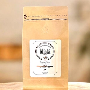 Może przedstawiać: Brązowa papierowa torba z zamkiem błyskawicznym. Torba ma białą etykietę z tekstem "Miski Coffee" i "Peruvian Primor". Etykieta zawiera r&oacute;wnież wskaźnik stopnia palenia z paskiem pokazującym stopień palenia jako "Średni".