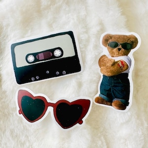 Puede incluir: Tres pegatinas que muestran un casete negro y beige, un oso de peluche marr&oacute;n con gafas de sol negras, camiseta blanca y vaqueros azules con un tatuaje de coraz&oacute;n en el brazo, y unas gafas de sol rojas con forma de coraz&oacute;n y lentes oscuras.