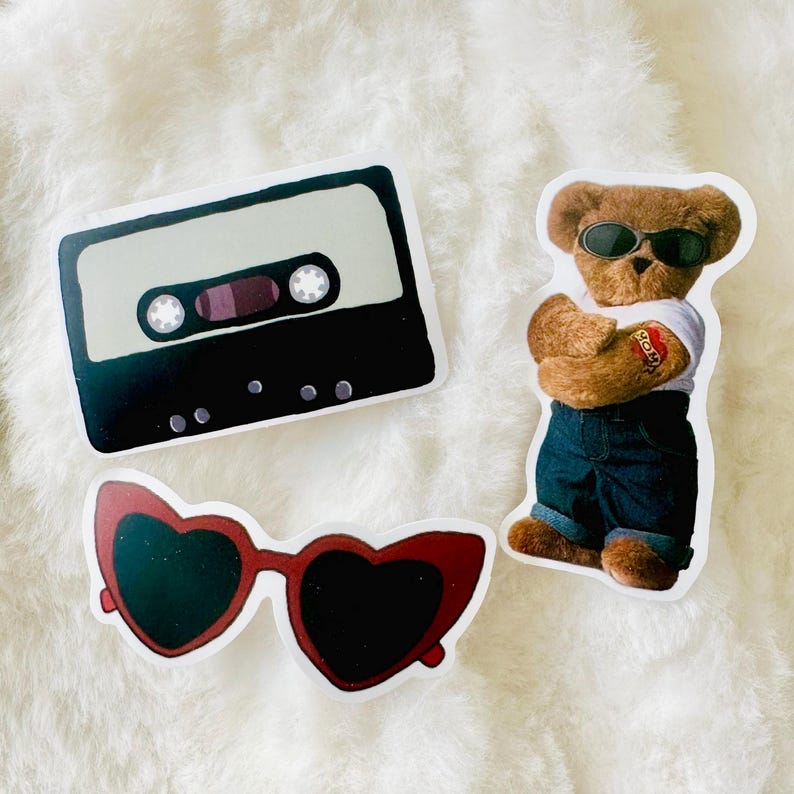 Puede incluir: Tres pegatinas que muestran un casete negro y beige, un oso de peluche marr&oacute;n con gafas de sol negras, camiseta blanca y vaqueros azules con un tatuaje de coraz&oacute;n en el brazo, y unas gafas de sol rojas con forma de coraz&oacute;n y lentes oscuras.