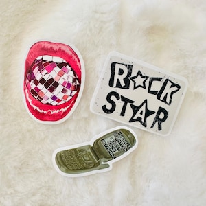 May include: Three stickers: one featuring a pair of bright red lips biting into a multi-colored disco ball, a rectangular sticker with the black text ROCK STAR, and a green flip phone sticker with the text I'M ONLY SEVENTEEN I DON'T KNOW ANYTHING.