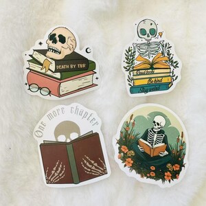 May include: Four stickers featuring skeletons reading books. The stickers are white with a rounded rectangular shape. The first sticker features a skull with a stack of books that say "Death by TBR". The second sticker features a skeleton sitting on a stack of books that say "Read books, Be kind, Stay weird". The third sticker features a skeleton reading a book with the text "One more chapter". The fourth sticker features a skeleton sitting in a field of flowers reading a book.