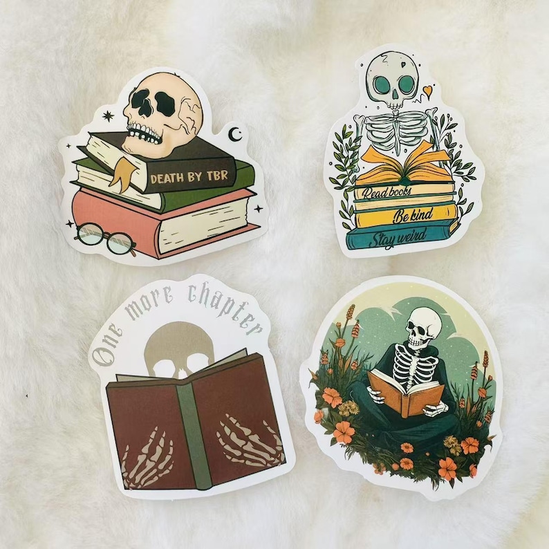 May include: Four stickers featuring skeletons reading books. The stickers are white with a rounded rectangular shape. The first sticker features a skull with a stack of books that say "Death by TBR". The second sticker features a skeleton sitting on a stack of books that say "Read books, Be kind, Stay weird". The third sticker features a skeleton reading a book with the text "One more chapter". The fourth sticker features a skeleton sitting in a field of flowers reading a book.