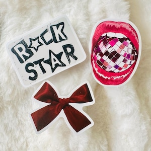 Puede incluir: Tres pegatinas que incluyen una pegatina rectangular blanca con el texto negro ROCK STAR, una pegatina de una boca abierta con labios rojos brillantes que contiene una bola de discoteca multicolor y una pegatina de un lazo de terciopelo rojo oscuro.