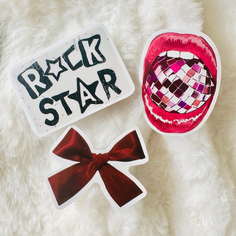 Puede incluir: Tres pegatinas que incluyen una pegatina rectangular blanca con el texto negro ROCK STAR, una pegatina de una boca abierta con labios rojos brillantes que contiene una bola de discoteca multicolor y una pegatina de un lazo de terciopelo rojo oscuro.