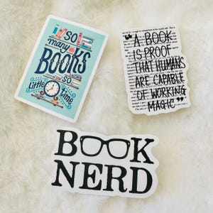 Peut inclure: Trois autocollants rectangulaires et d&eacute;coup&eacute;s. Un autocollant bleu clair pr&eacute;sente des livres color&eacute;s et le texte 'so many BOOKS so little time'. Un autocollant blanc affiche le texte noir 'A BOOK IS PROOF THAT HUMANS ARE CAPABLE OF WORKING MAGIC'. Un autocollant blanc indique 'BOOK NERD' avec des lunettes.