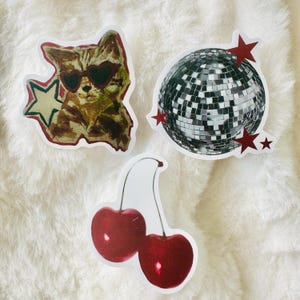 Puede incluir: Tres pegatinas troqueladas que muestran un gato marr&oacute;n con gafas de sol en forma de coraz&oacute;n junto a una estrella verde, una bola de discoteca plateada con estrellas rojas y dos cerezas rojas brillantes con un tallo verde sobre fondo blanco.