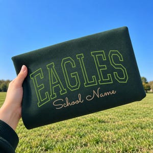 May include: A dark green sweatshirt featuring the word EAGLES in a lime green collegiate font, with the text School Name written below in a light peach cursive script. The garment is held up against a clear blue sky and a grassy field.