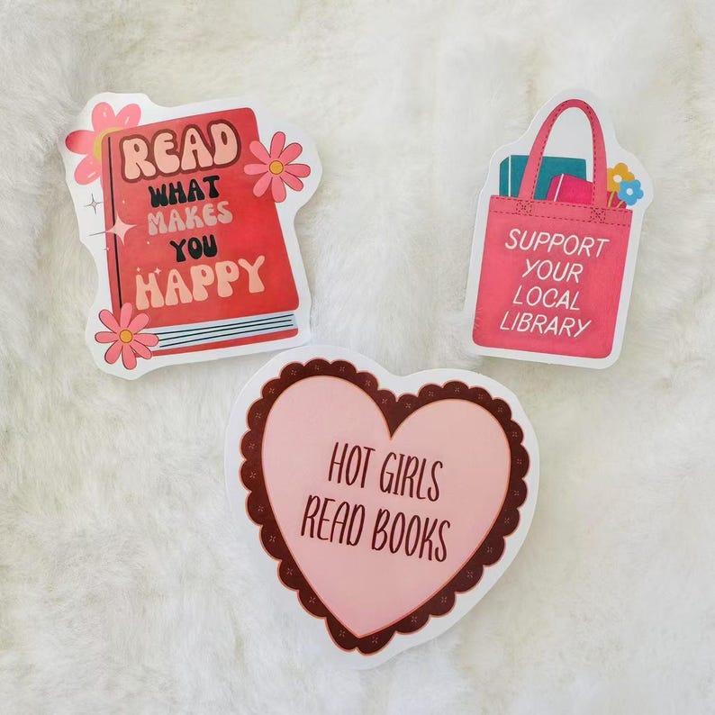 May include: Three pink and red stickers with different messages. The first sticker features a red book with the text "Read What Makes You Happy". The second sticker is a pink tote bag with the text "Support Your Local Library". The third sticker is a pink heart with the text "Hot Girls Read Books".
