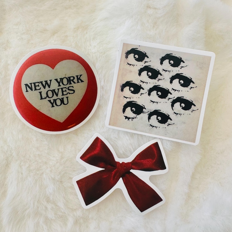 Może przedstawiać: Trzy naklejki: okrągła czerwona naklejka z kremowym sercem zawierającym czarny napis NEW YORK LOVES YOU, kwadratowa naklejka z kremowym tłem przedstawiająca wiele czarnych ilustrowanych oczu oraz wycinana naklejka w kształcie ciemnoczerwonej satynowej kokardki.