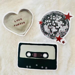 Może przedstawiać: Trzy naklejki przedstawiające beżowy obiekt w kształcie serca z napisem LOVE ALWAYS, srebrną kulę dyskotekową z czerwonymi gwiazdkami oraz czarno-beżową kasetę magnetofonową z dwiema okrągłymi szpulami i prostokątnym okienkiem pośrodku.