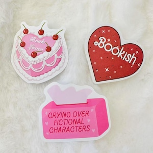 May include: Three pink and white stickers with bookish themes. The first sticker features a pink cake with cherries and the text "Book Lover". The second sticker is a red heart with the text "Bookish". The third sticker is a pink tissue box with the text "Crying Over Fictional Characters".