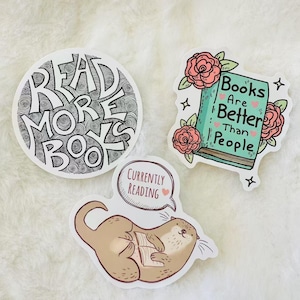 Peut inclure: Trois autocollants en vinyle avec des motifs sur le th&egrave;me des livres. Un autocollant circulaire porte l'inscription READ MORE BOOKS en noir. Un deuxi&egrave;me montre un livre bleu sarcelle avec des roses et le texte Books Are Better Than People.