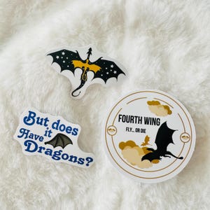 May include: Three stickers featuring dragons. One sticker has a black dragon with yellow accents and stars on its wings. Another sticker has a black dragon with a white background and the text "Fourth Wing Fly... or Die". The third sticker has a black dragon with blue text that reads "But does have it Dragons?".