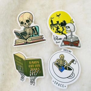 May include: Four skeleton themed stickers with different designs. One sticker features a skeleton reading a book with the text "Read Banned Books". Another sticker features a skeleton reading a book with the text "Hands Off Our Books". A third sticker features a skeleton sitting in a cup with the text "I Need My Space". The final sticker features a skeleton reading a book with the text "Welcome Home".