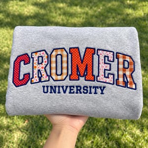 May include: A light gray sweatshirt featuring the text CROMER UNIVERSITY embroidered in a collegiate font. The letters of CROMER are filled with various patterns including red polka dots, floral prints, and orange gingham checks.