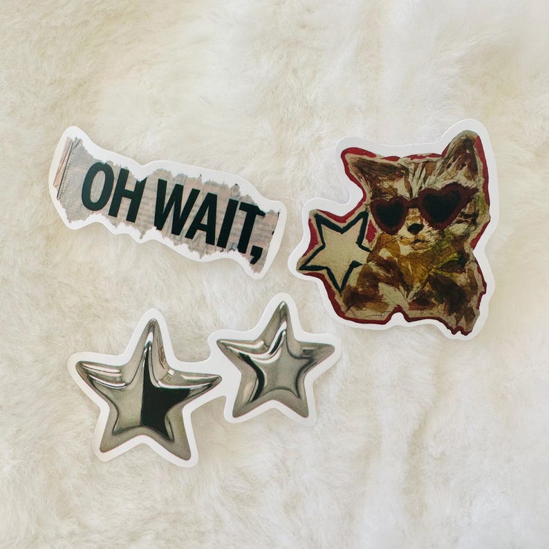May include: A set of four stickers including one with the text OH WAIT, a sticker of a cat wearing heart-shaped sunglasses next to a star, and two individual silver-colored star stickers with a metallic finish and a white border.