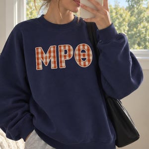 May include: A navy blue crewneck sweatshirt featuring the letters MPO in a white and orange gingham pattern. The letters are applied as large, stitched-on patches across the chest. The sweatshirt has long sleeves with ribbed cuffs and a ribbed neckline.