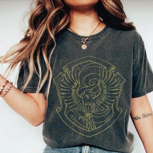May include: Dark gray t-shirt featuring a yellow crest with a stylized eagle design. The shirt has a vintage, worn look. The design is centered on the front of the shirt, creating a bold statement.