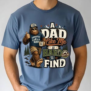 May include: A dusty blue t-shirt featuring a graphic of a brown Bigfoot character wearing a cap, sunglasses, and a blue shirt, holding a coffee mug. The text reads A DAD Like Me is HARD To FIND, with a small badge stating HAPPY FATHER'S DAY.