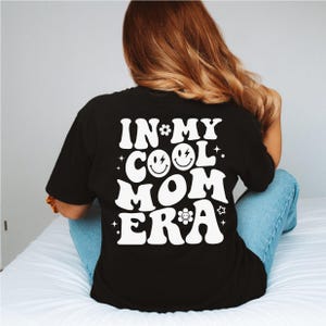 a woman sitting on a bed wearing a black shirt that says in my cool mom