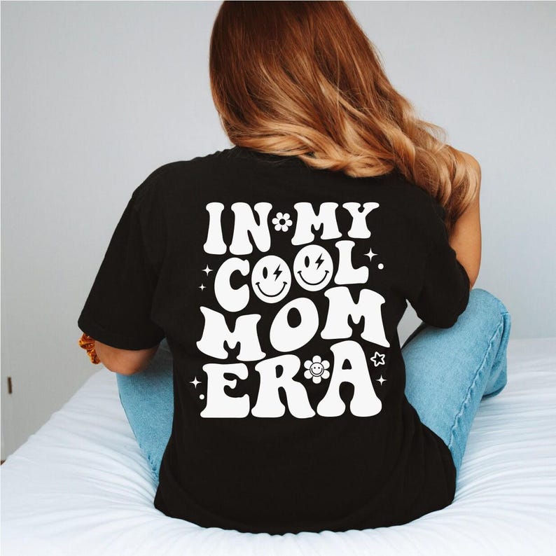 a woman sitting on a bed wearing a black shirt that says in my cool mom