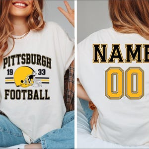 May include: White t-shirt with black and yellow text and a football helmet graphic. The front of the shirt says "Pittsburgh Football" with the numbers "19" and "33" on either side of the helmet. The back of the shirt says "NAME" above the number "00".